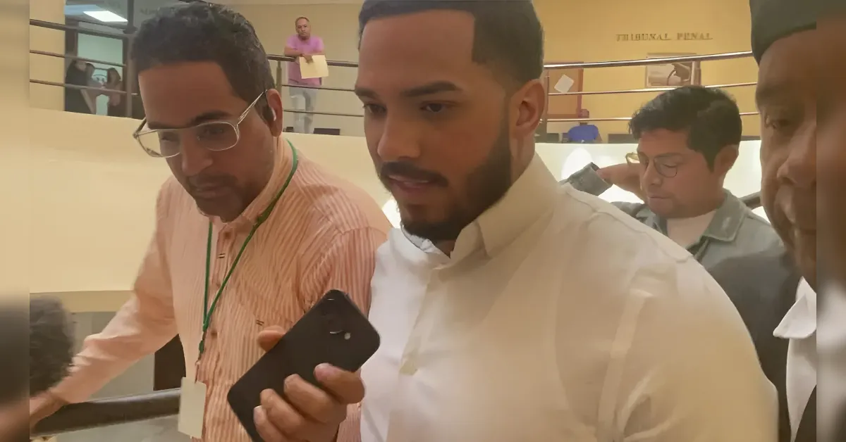 Man in a white shirt holding a black smartphone while two colleagues stand nearby in a lobby or hallway setting.