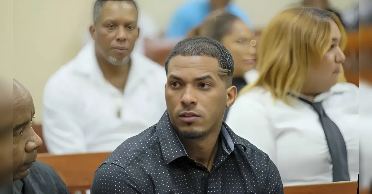 Young man with a short haircut and goatee wearing a dark dotted shirt sits in a courtroom bench, with other attendees in the background .