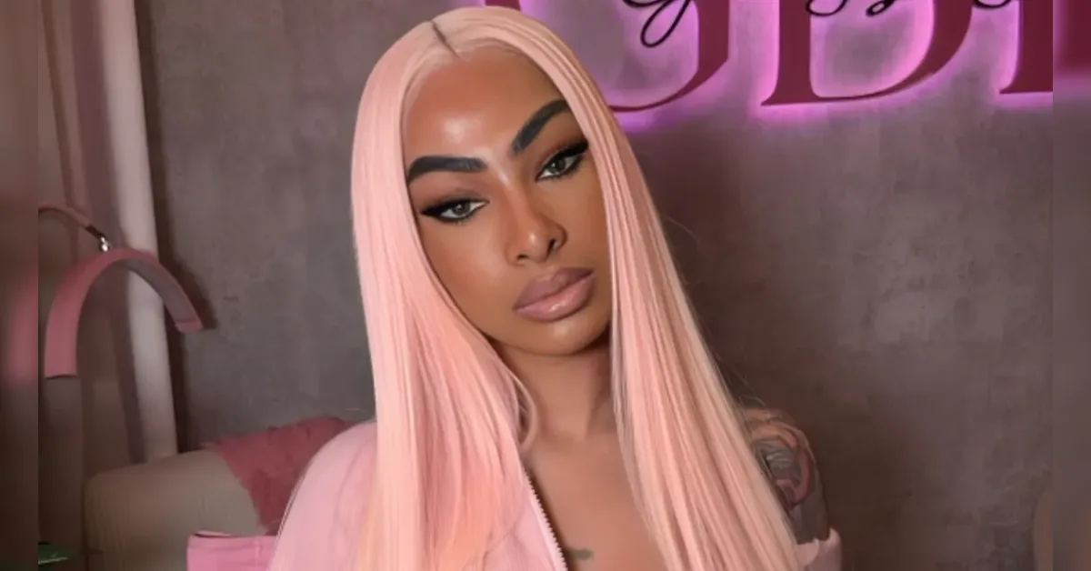 Person with long, straight pastel pink hair and bold makeup posing indoors against a pink neon backdrop.