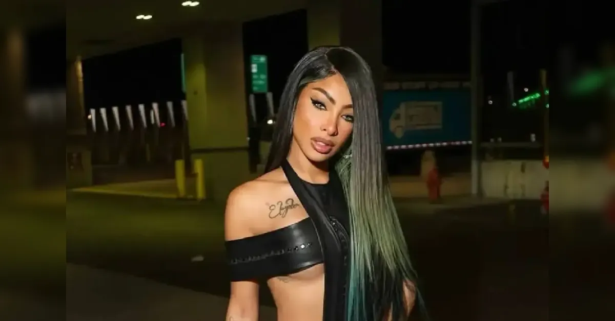 Portrait of a woman with long dark hair and green tips, wearing a black off‑the‑shoulder top, posing outdoors at night in an urban area, with tattoos on her left shoulder plainer.
