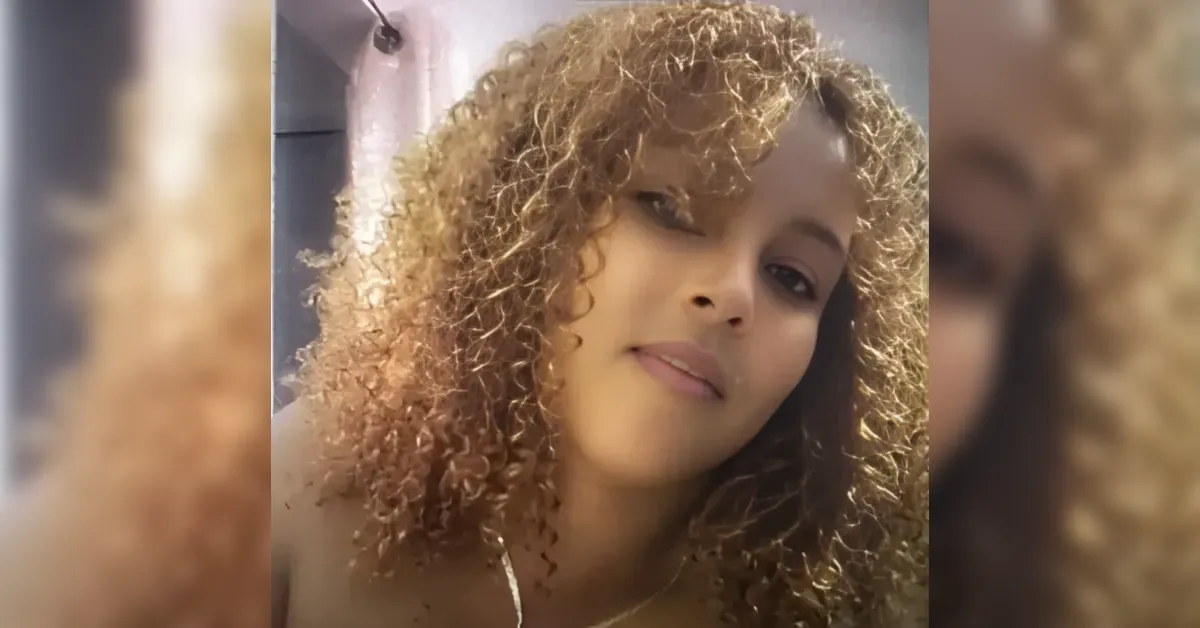 Close-up selfie of a woman with shoulder-length, tight curls looking at the camera with a gentle smile indoors