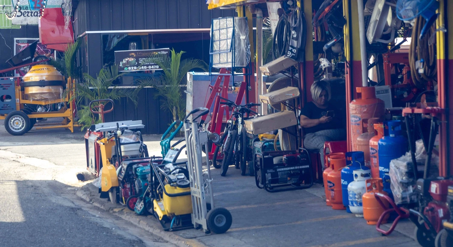 Outdoor workshop with stacked tools, bikes, and gas cylinders; a person sits inside on the right using a phone.