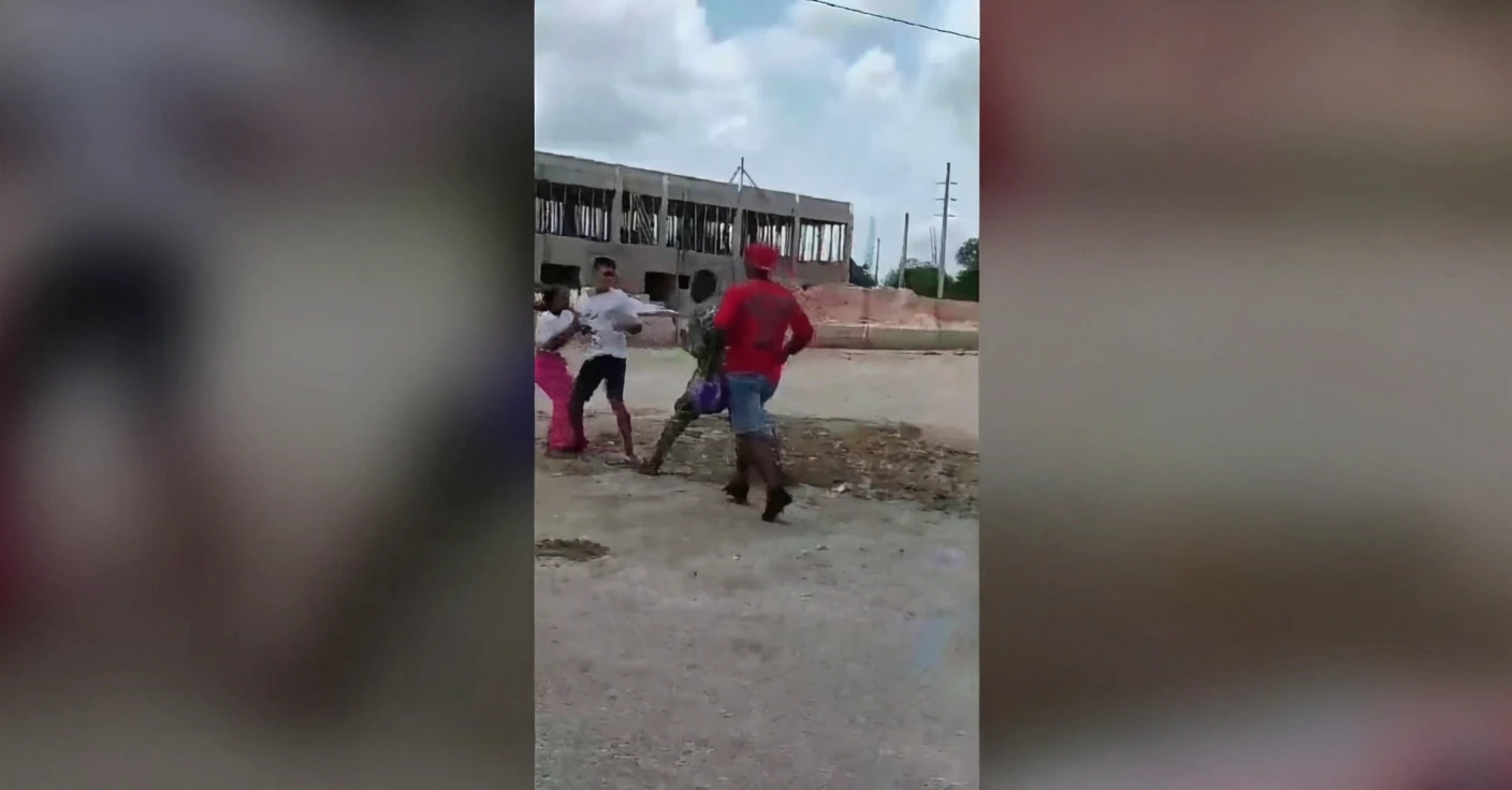 Several people argue and push each other on a dusty construction site, with a half-built building in the background.
