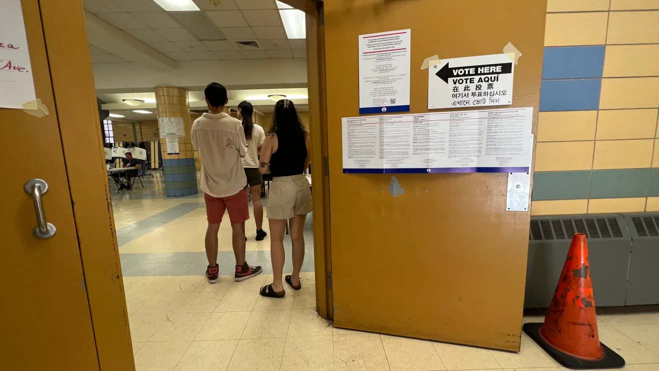 Three people stand with their backs to the camera in a school hallway near a bulletin board and a 'Vote Here' sign; orange doors and a traffic cone nearby.