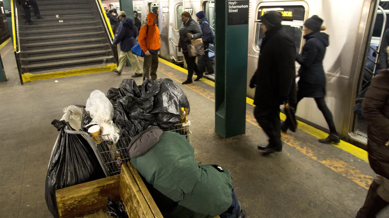 Cart overflowing with trash bags and items sits on a subway platform as commuters walk by and a train doors are open nearby.