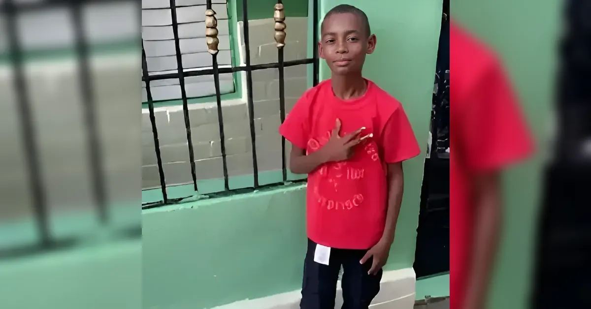 Boy in a red t-shirt stands in front of a green wall and black metal gate, hand over his chest as if pledging or saluting.