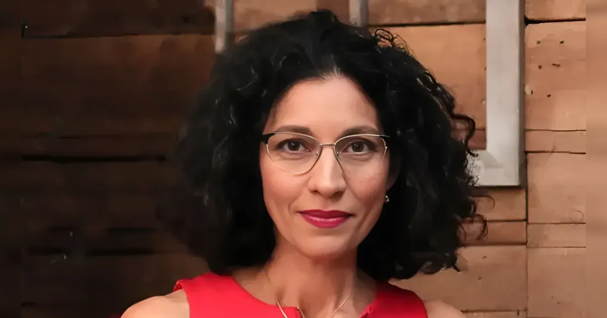 Portrait of a woman with curly black hair, wearing glasses and a red top, smiling in front of a wooden background.