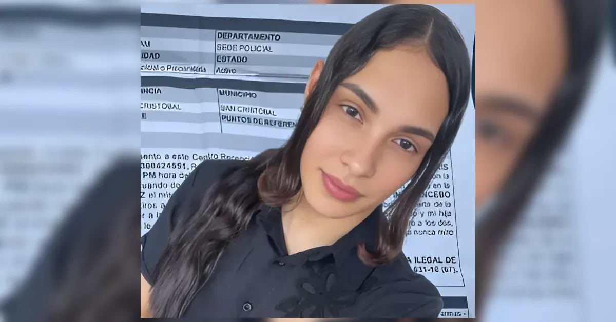 Portrait of a young woman with long dark hair wearing a black shirt, taking a selfie in front of a striped backdrop that resembles a mugshot board in a bright setting.