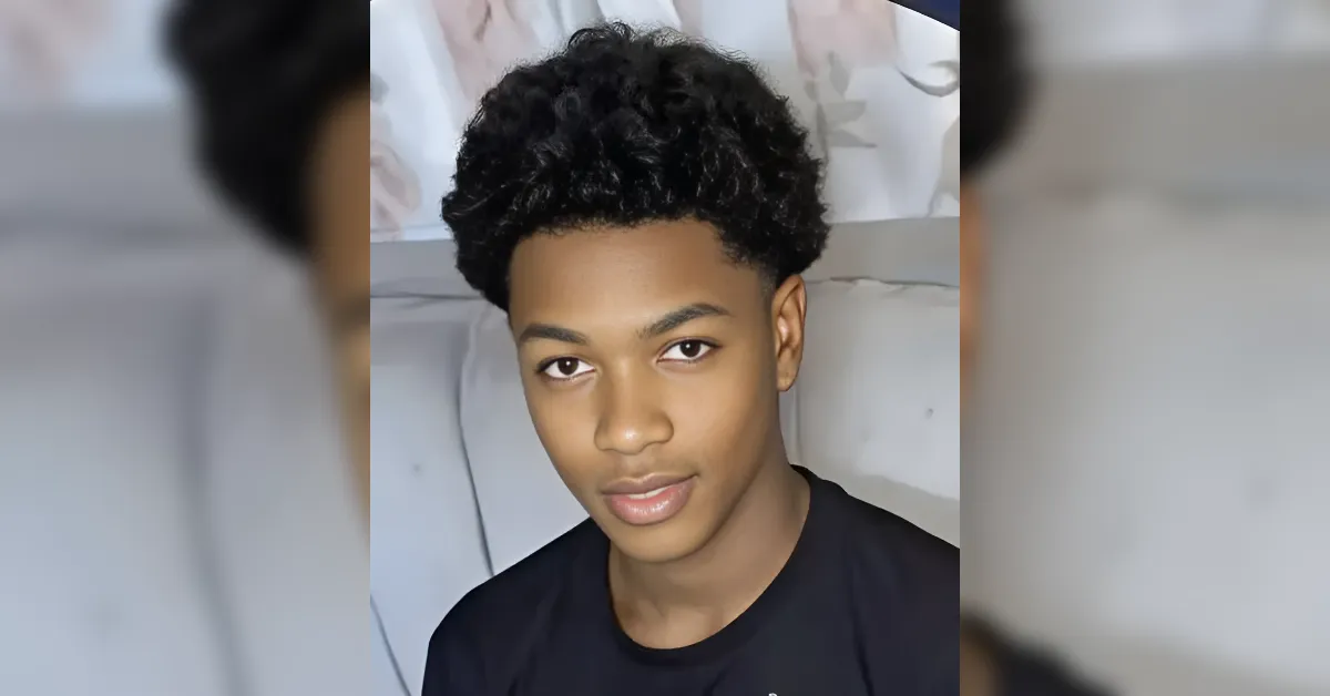 Young man with short curly black hair looking at the camera, wearing a dark shirt indoors against a neutral background.
