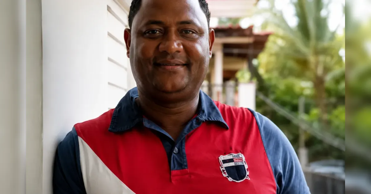 Smiling man leaning against an outdoor wall in a red and blue polo shirt with a crest on the chest area.