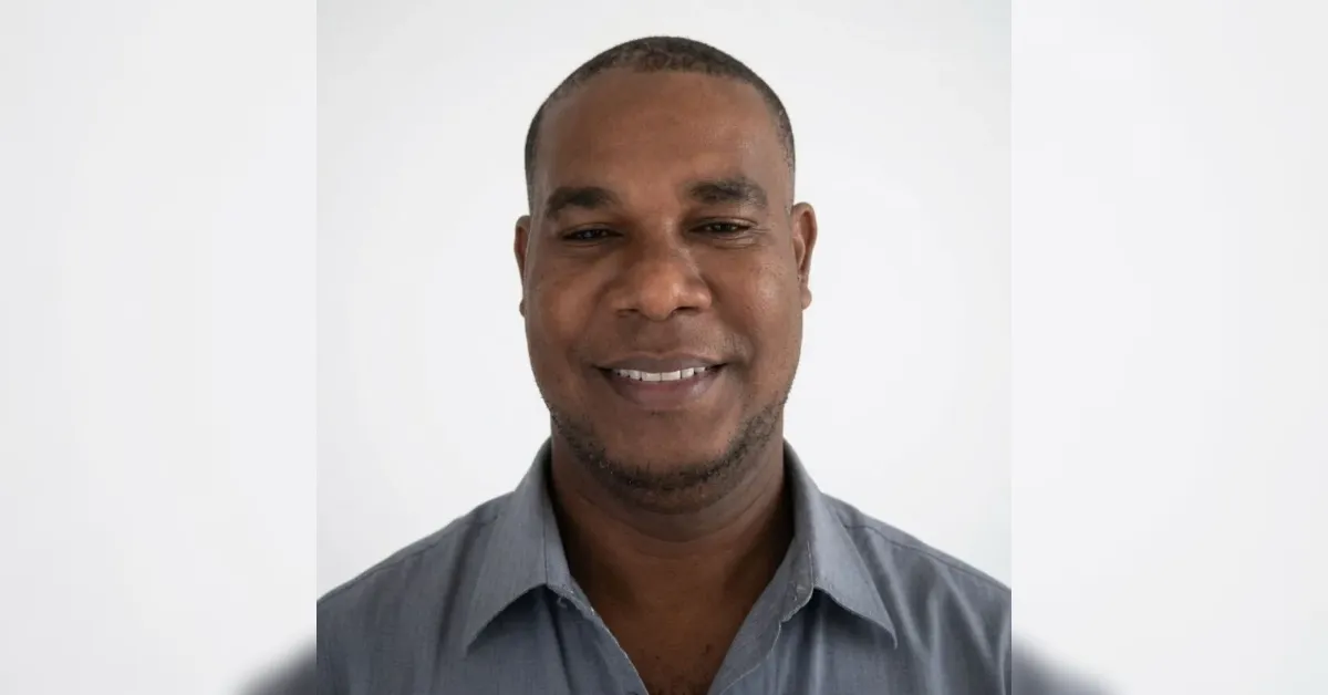 Man wearing a gray collared shirt smiles at the camera against a plain white background.