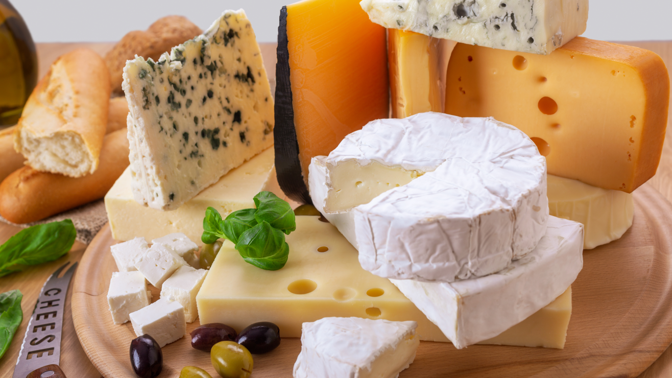 Assorted cheeses (brie, blue, Swiss, cheddar) on a wooden board with olives, basil, and bread slices.
