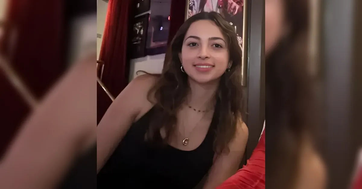 Smiling young woman with wavy brown hair in a black sleeveless top, wearing a pendant necklace and hoop earrings in a warmly lit indoor setting.