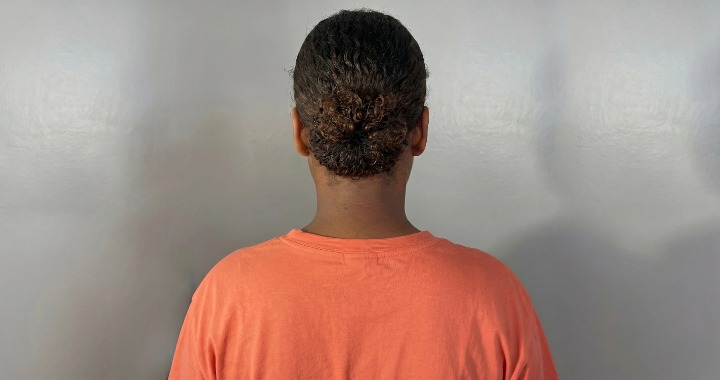 Rear view of a person with short curly hair tied in a bun, wearing an orange shirt against a gray wall.