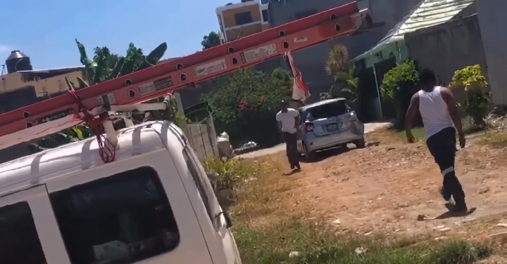 White utility truck with a long red ladder extended overhead; two men walk along a dirt road toward a small silver car in a residential area.