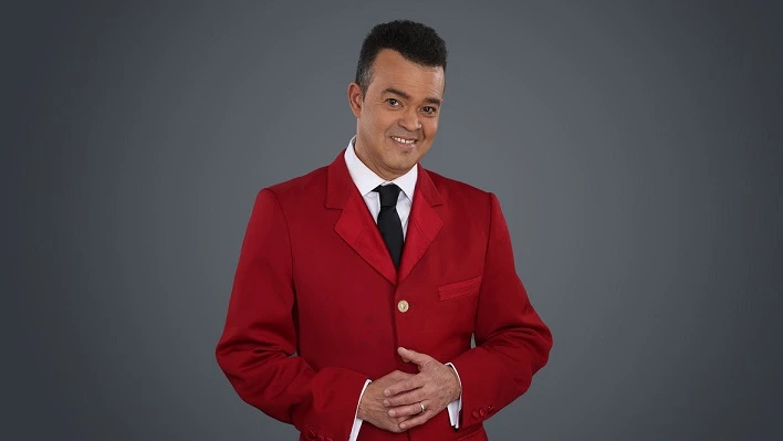 Portrait of a man in a red suit with a white shirt and black tie, smiling with hands clasped.