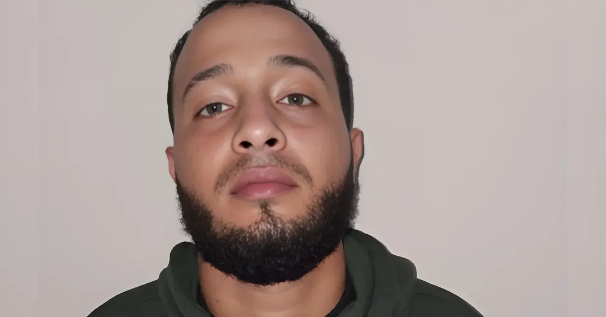 Close-up of a man with a full beard and short hair, wearing a dark green hoodie, looking at the camera against a plain wall.
