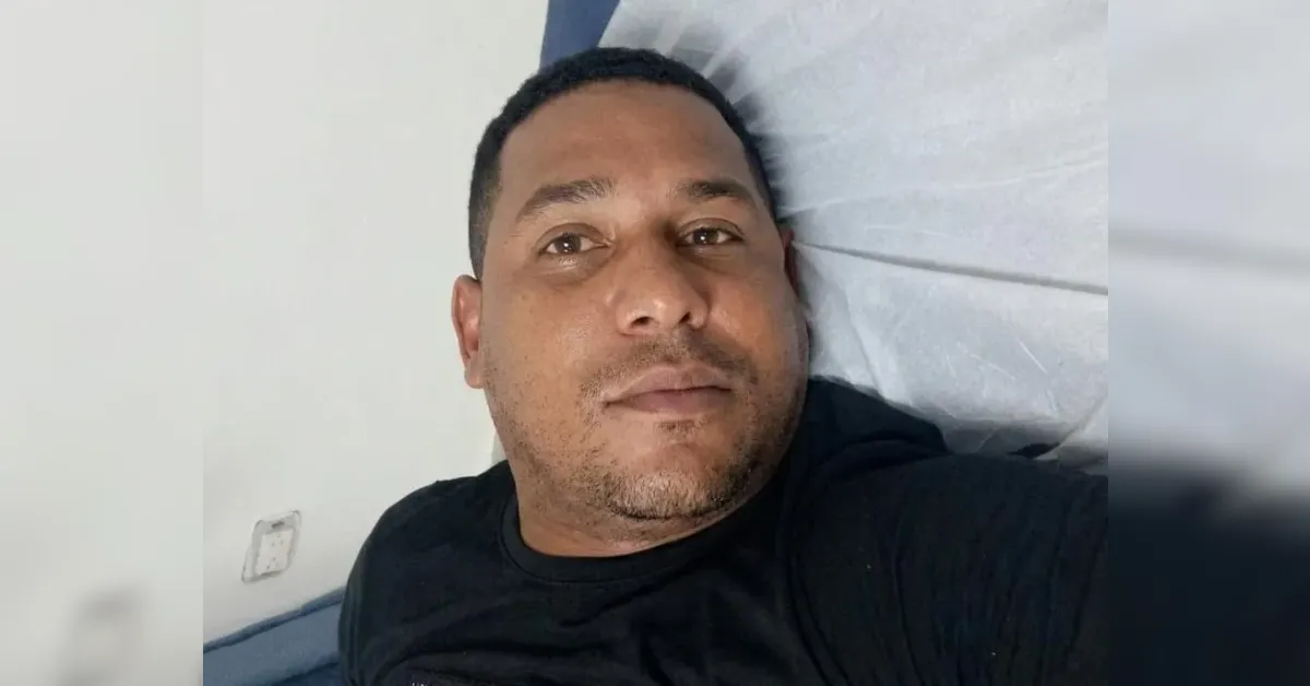 Man lying on a bed or couch, wearing a black shirt, facing the camera with a calm expression.