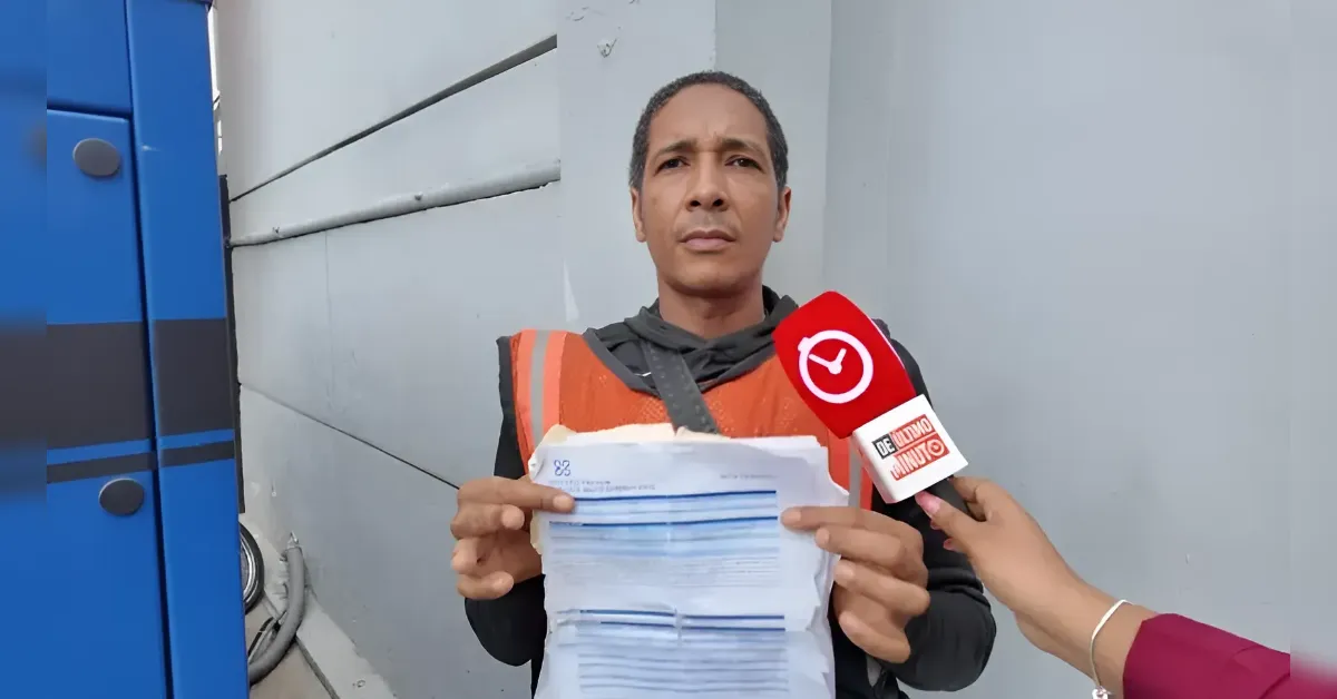 A man in an orange safety vest being interviewed, holding a stack of papers as a reporter with a red microphone speaks to him outside a gray wall and blue equipment.