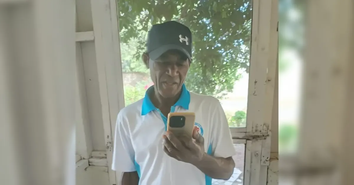 Older man wearing a black cap and white polo with teal collar, looking at a smartphone in a doorway with greenery outside.