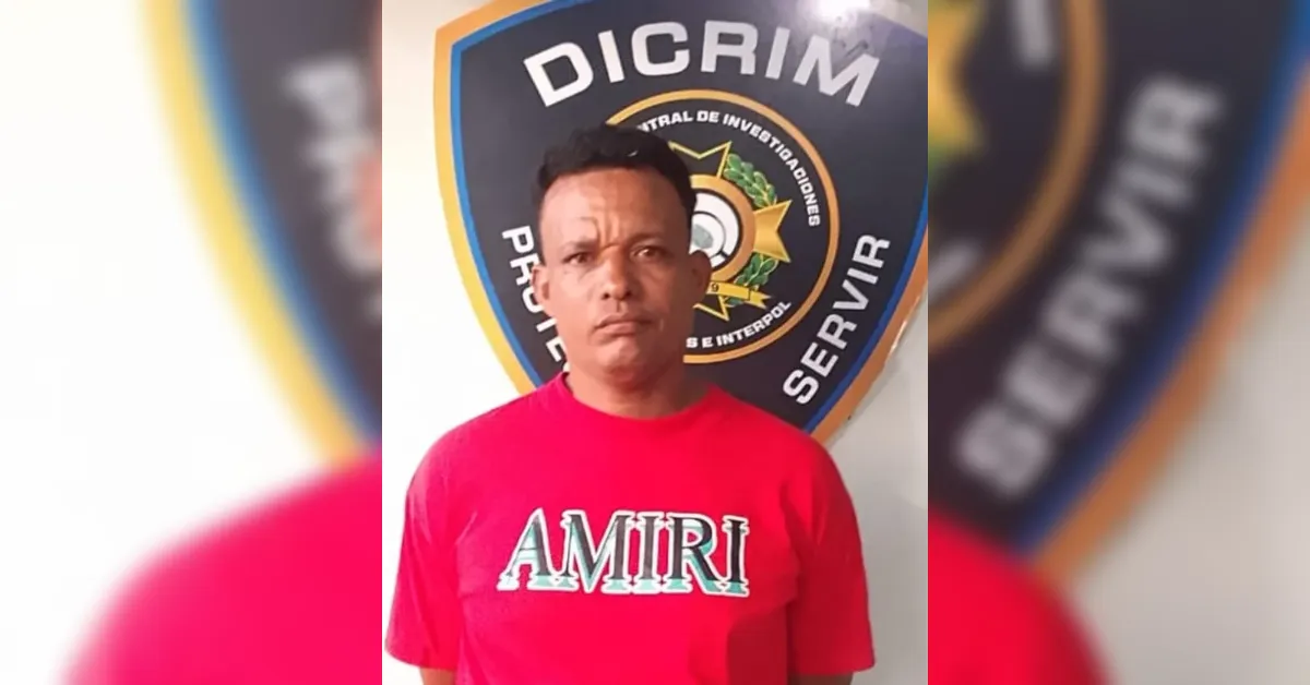 Man wearing a bright red AMIRI t-shirt stands in front of a police emblem on the wall behind him.