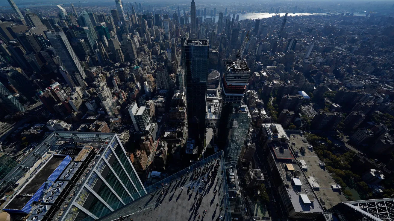 Aerial view of a dense city with skyscrapers and a glass-front viewing deck at the edge, with people looking down on the platform.