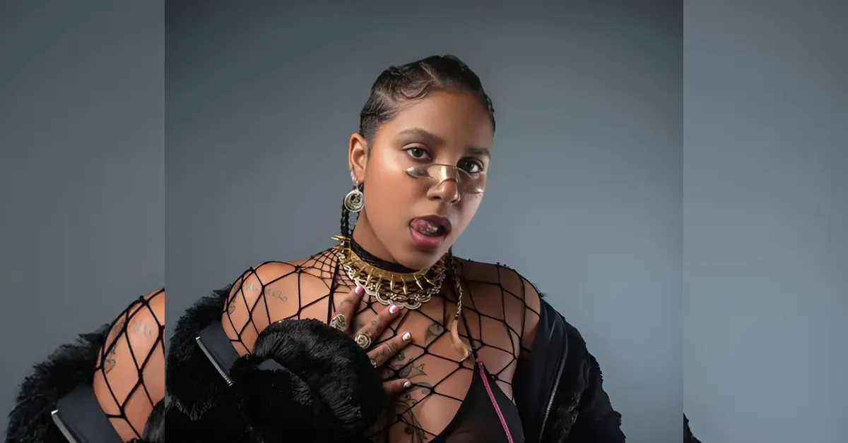 Woman with braided hair wearing a fishnet top and fur jacket, accessorized with gold jewelry, sticking her tongue out.
