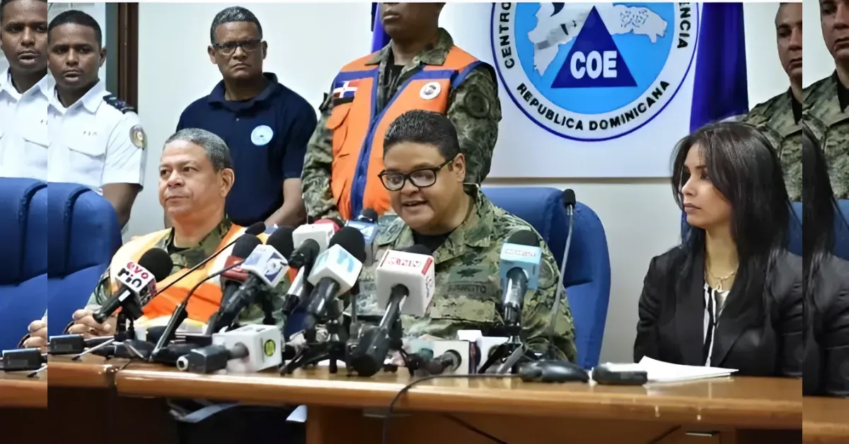 Military official in uniform speaks at a press conference with multiple microphones on the table, others seated behind him in a briefing room with a COE banner in the background.