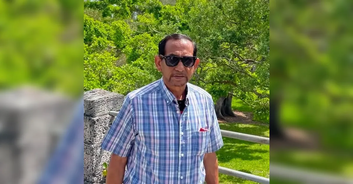 Man wearing sunglasses and a blue plaid short-sleeve shirt standing outdoors near a railing with green trees in the background.