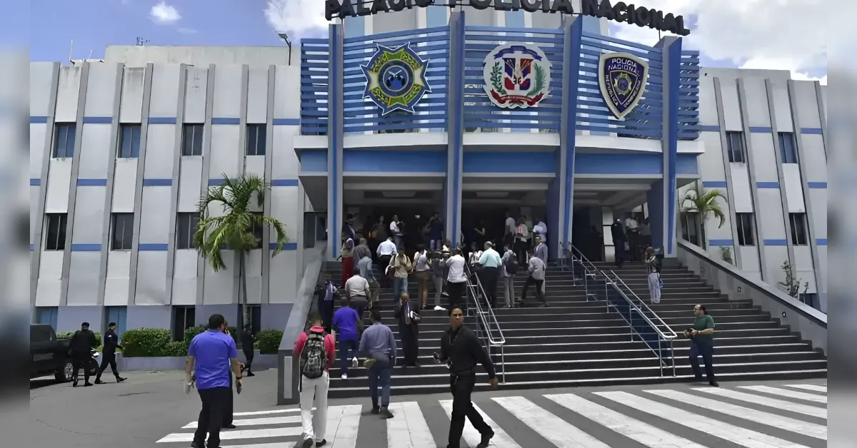 Front entrance of a police station with blue and white façade and three crest emblems, as people queue up the stairs to enter.