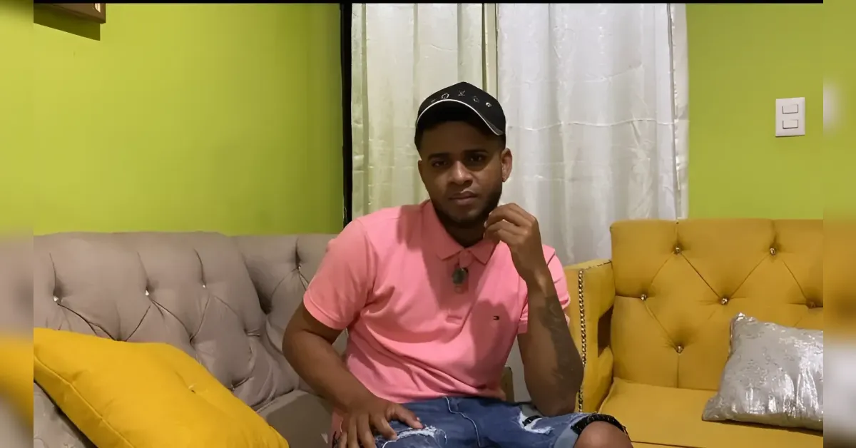Young man in a pink polo shirt and black cap sits on a beige tufted sofa with yellow cushions in a lime-green living room, looking at the camera.