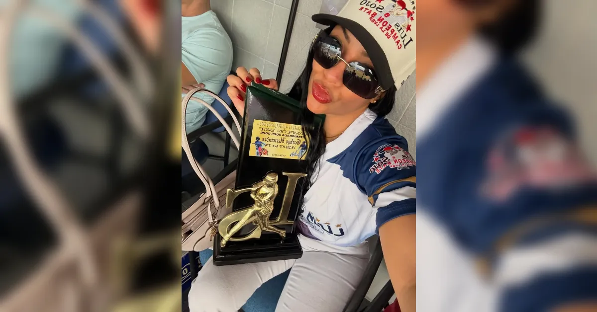 Woman wearing a cap and sunglasses holding a black trophy case with a gold baseball figure and a yellow label, smiling for the camera.