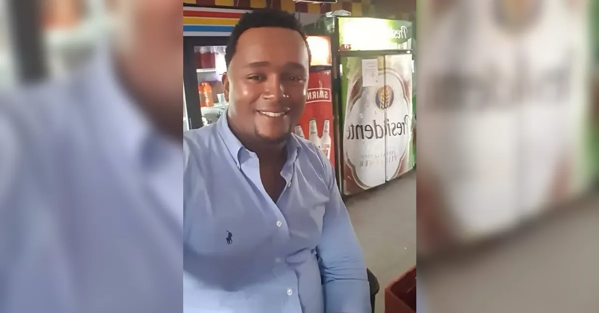 Smiling man in a light blue button-down shirt sits at a store counter with soda fridges in the background.