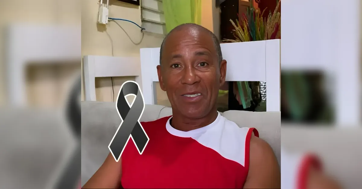 Smiling man in a red and white sleeveless shirt sits on a couch indoors, with a black awareness ribbon graphic over his chest.