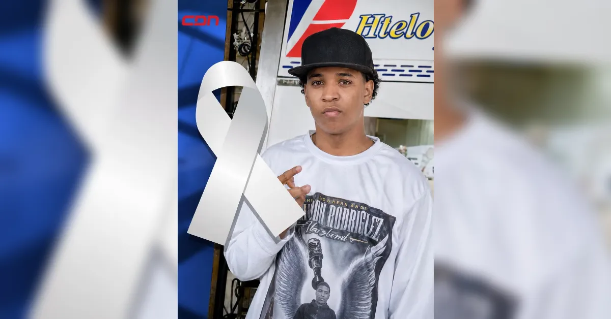 Young man in a white graphic shirt and black cap posing with a large white awareness ribbon in front of a blue backdrop.