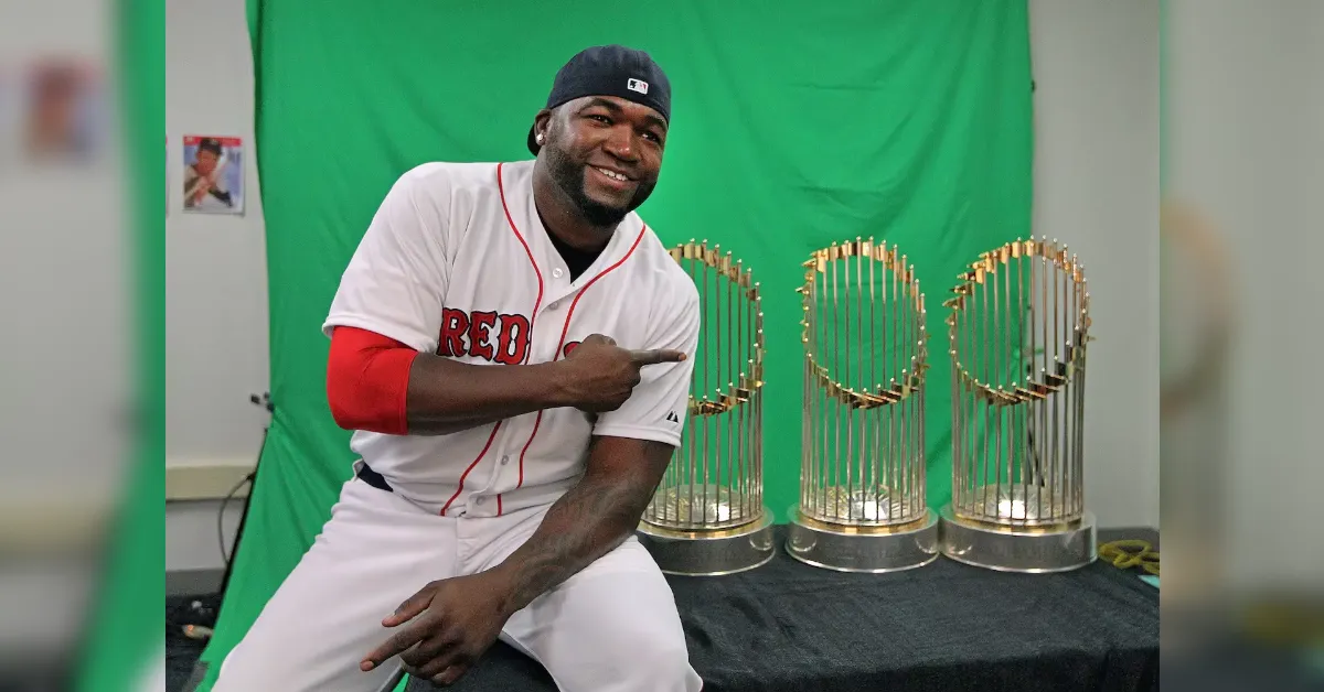 Smiling man in a baseball jersey points to three gold trophies on a table against a green screen backdrop.