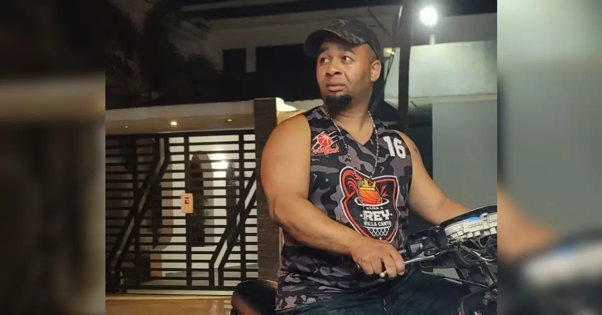 Man in a camo sleeveless shirt and cap sits on a motorcycle at night, looking to the side with a puzzled expression.