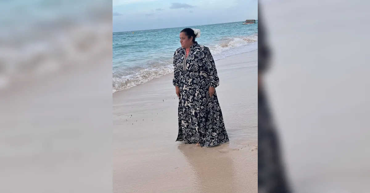 Woman stands in shallow water on a beach, wearing a long black-and-white floral dress with a white flower in her hair, looking toward the sea.