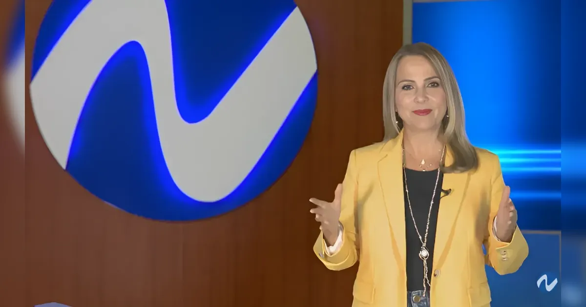Female news anchor wearing a yellow blazer stands in a studio with a blue backdrop and a large circular logo behind her.