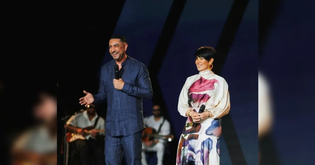 Man in blue shirt speaks into a microphone on stage, gesturing with his other hand, while a woman in a colorful dress stands beside him smiling.