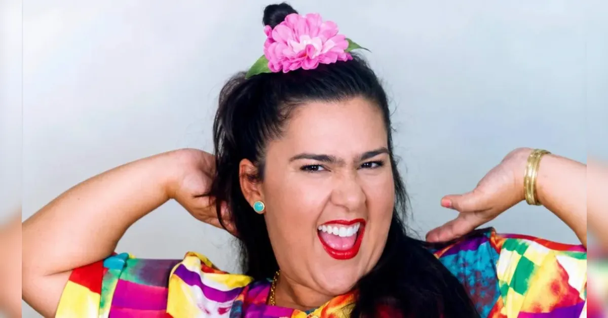 Joyful woman with a pink flower in her dark hair, bright colorful dress, turquoise earrings, and red lipstick, posing with hands behind her head.
