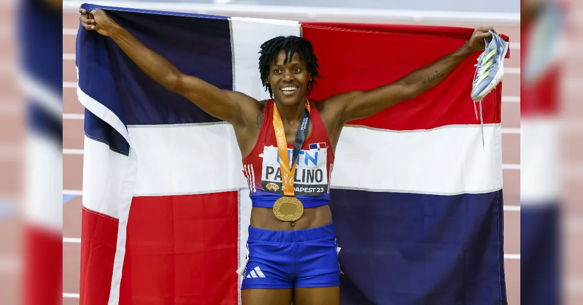 Female sprinter with a gold medal, arms raised in victory, standing in front of a red, white, and blue flag.