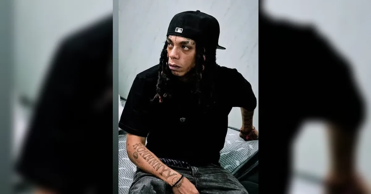 Young man with braids wearing a black cap and t-shirt sits on a patterned bed, looking off to the side.
