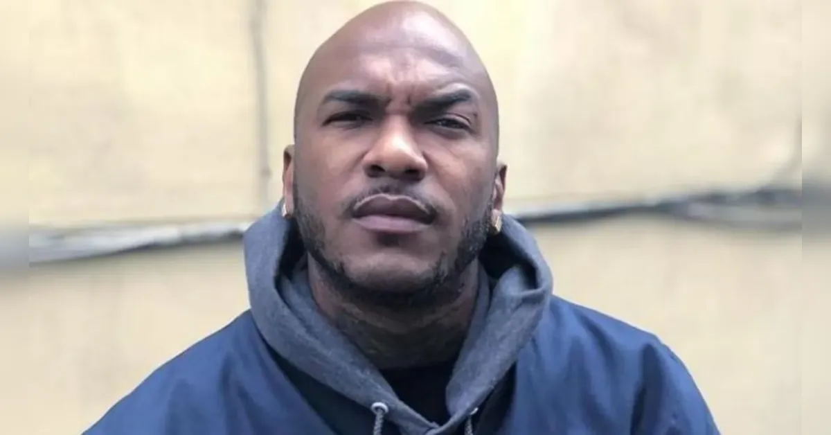 Man outdoors wearing a blue jacket and gray hoodie, looking straight at the camera with a serious expression.