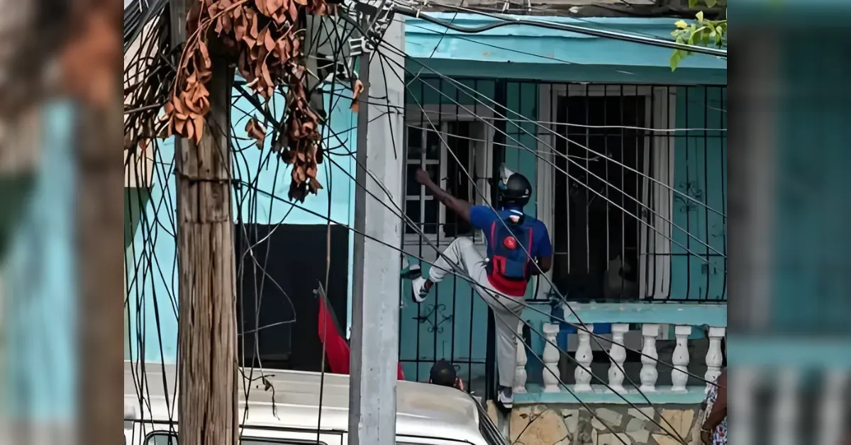 Man wearing a helmet and backpack on a turquoise house balcony, reaching toward a window amid tangled overhead wires.