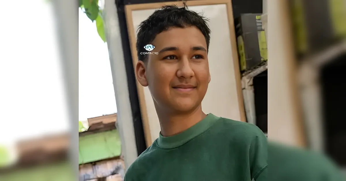 Young man with short black hair in a green shirt smiles while standing near a doorway outside/indoors.