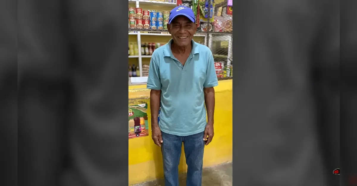 Smiling man in a light blue polo shirt and blue cap standing behind a yellow counter in a small shop.