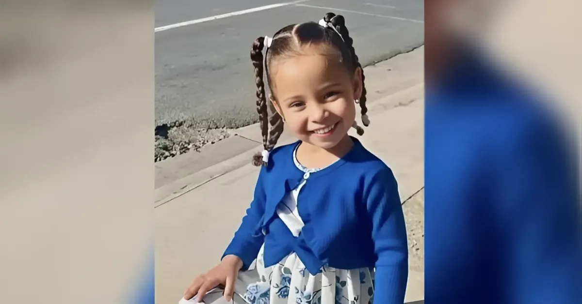Smiling young girl with braided pigtails wearing a blue cardigan and floral dress, standing on a sidewalk by the street.