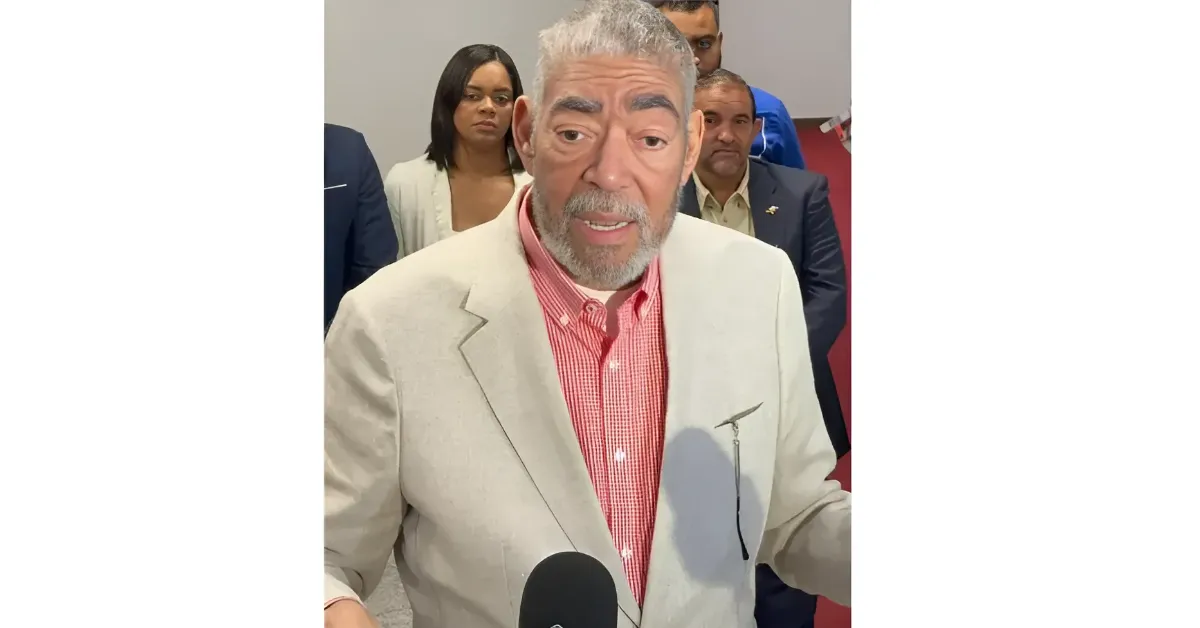 Older man with a gray beard in a light beige blazer and red checkered shirt speaking to reporters, microphone visible, with a group behind him in a hallway.