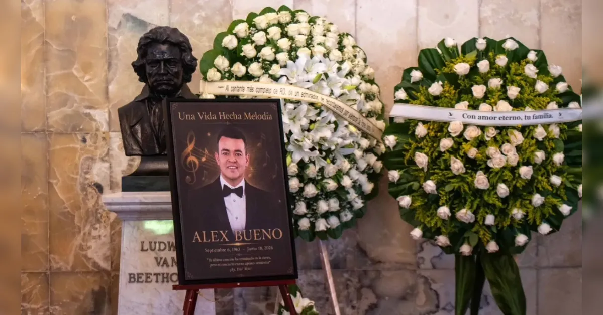 Memorial display with a framed portrait of Alex Bueno in a tuxedo, flanked by two large white flower wreaths.